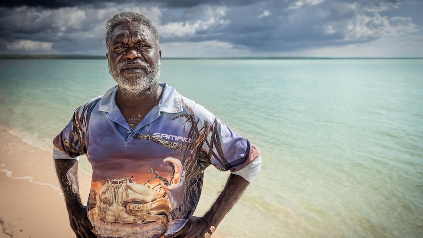 A man stands on a beach looking past the camera, his hands on his hips and storm clouds gathering behind him.