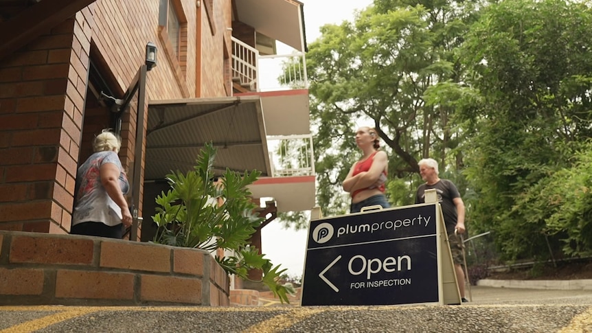 An open for inspection sign at a Brisbane apartment, behind it people are entering a building