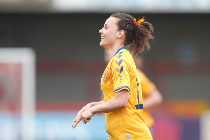 Hayley Raso smiles and holds her hands in front of her as seen from the side, wearing a yellow football kit