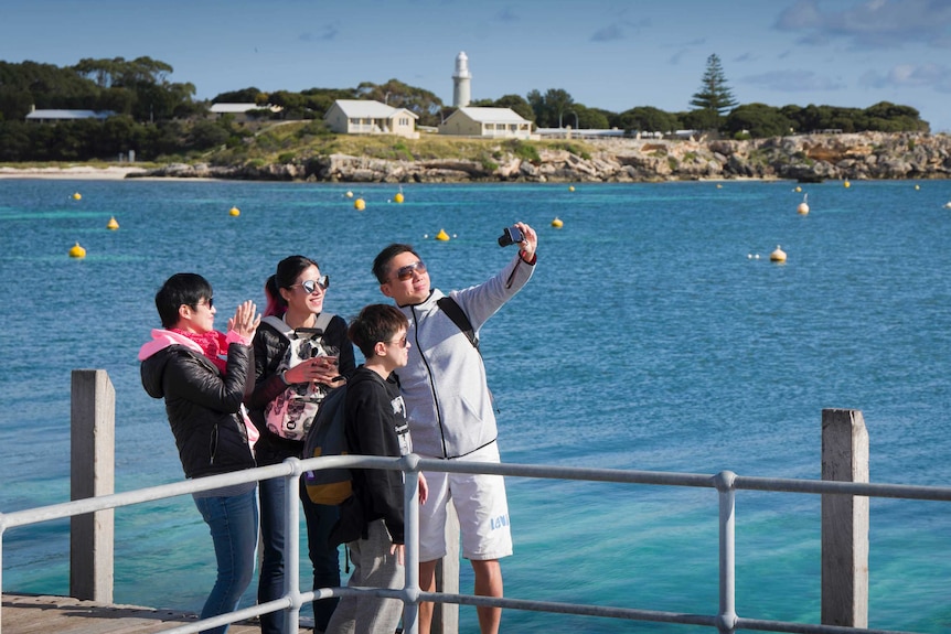 A group of four tourists stand on a pier taking a selfie on Rottnest Island with the ocean behind them.