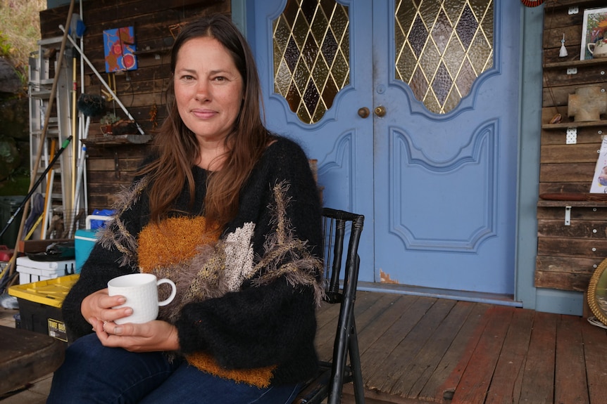 A woman in a fuzzy jumper sits holding a tea cup on a patio, with blue doors behind her.