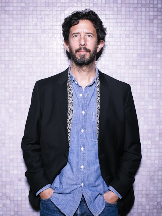 Middle-aged man with dark curly hair and beard wearing shirt and dark suit, against tiled backdrop.