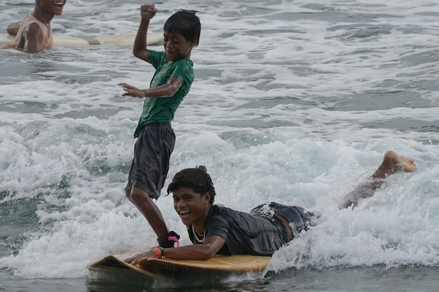 Two young boys in the white wash surf on boards smiling - one lying and one standing.
