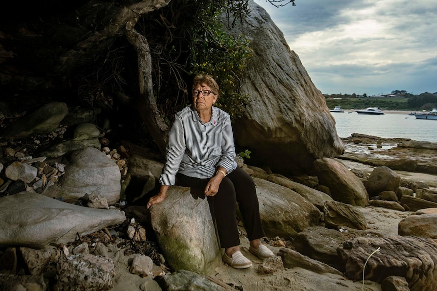 Aboriginal elder Aunty Barbara Simms sits on the rock at the edge of a dark cave with a quiet bay behind her.