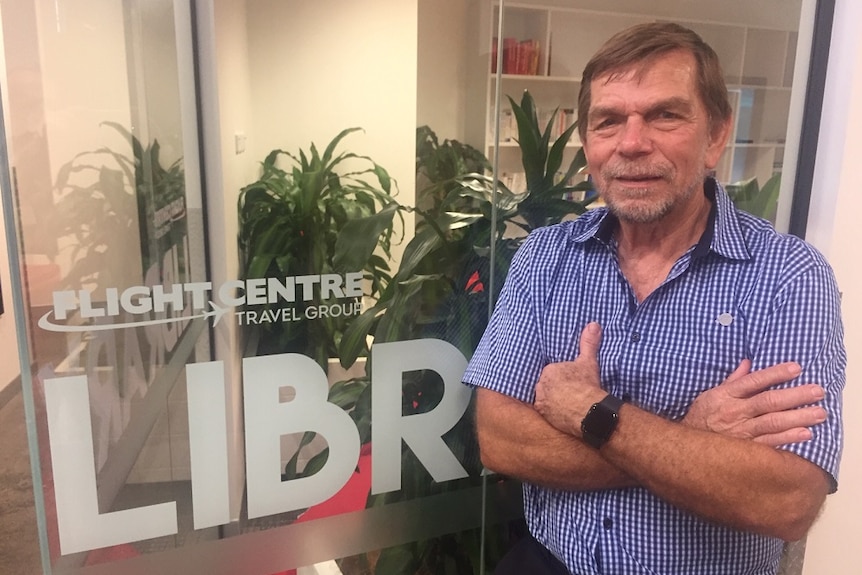Graham turner with his arms crossed looking at the camera and leaning against a glass office wall that says Flight Centre on it.