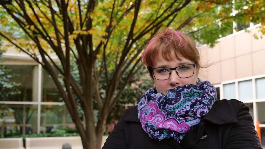 Carol is standing with her arms crossed to keep warm. She is wearing a black jacket and a colourful scarf hides half her face.