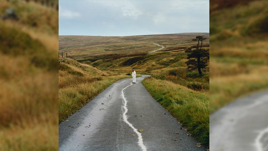 A man stands in the middle of an empty road in a deserted green field. He wears all white. 