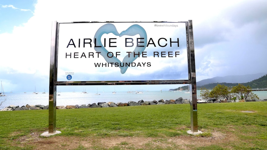 A sign on grass overlooking the ocean that says Airlie Beach, Heart of the Reef.