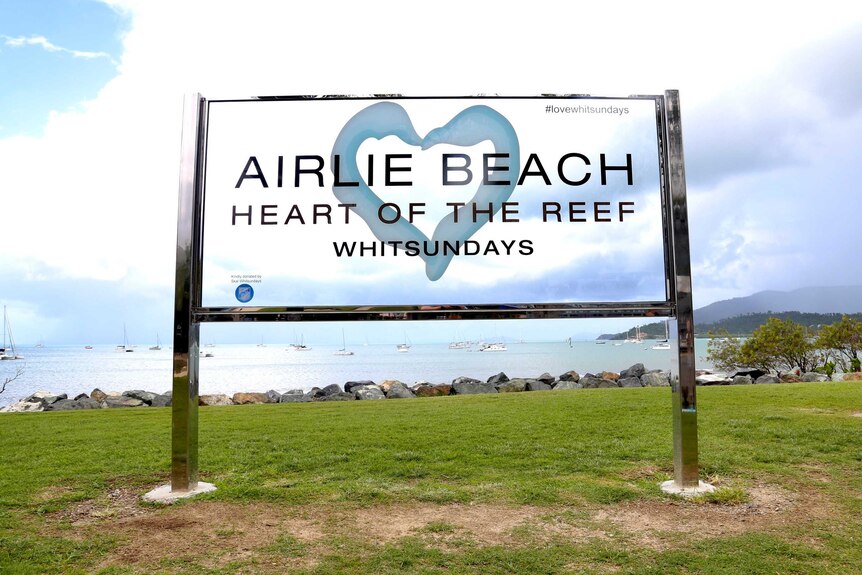 Un cartel en el césped con vistas al océano que dice Airlie Beach, Heart of the Reef.