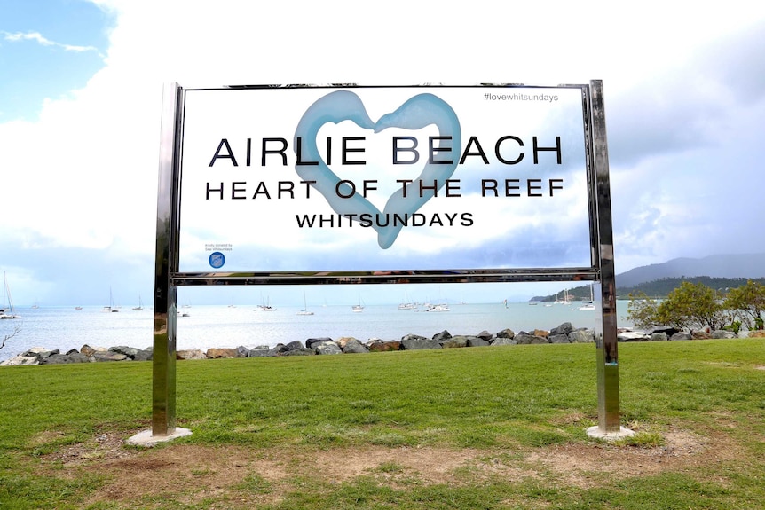 A sign on grass overlooking the ocean that says Airlie Beach, Heart of the Reef.