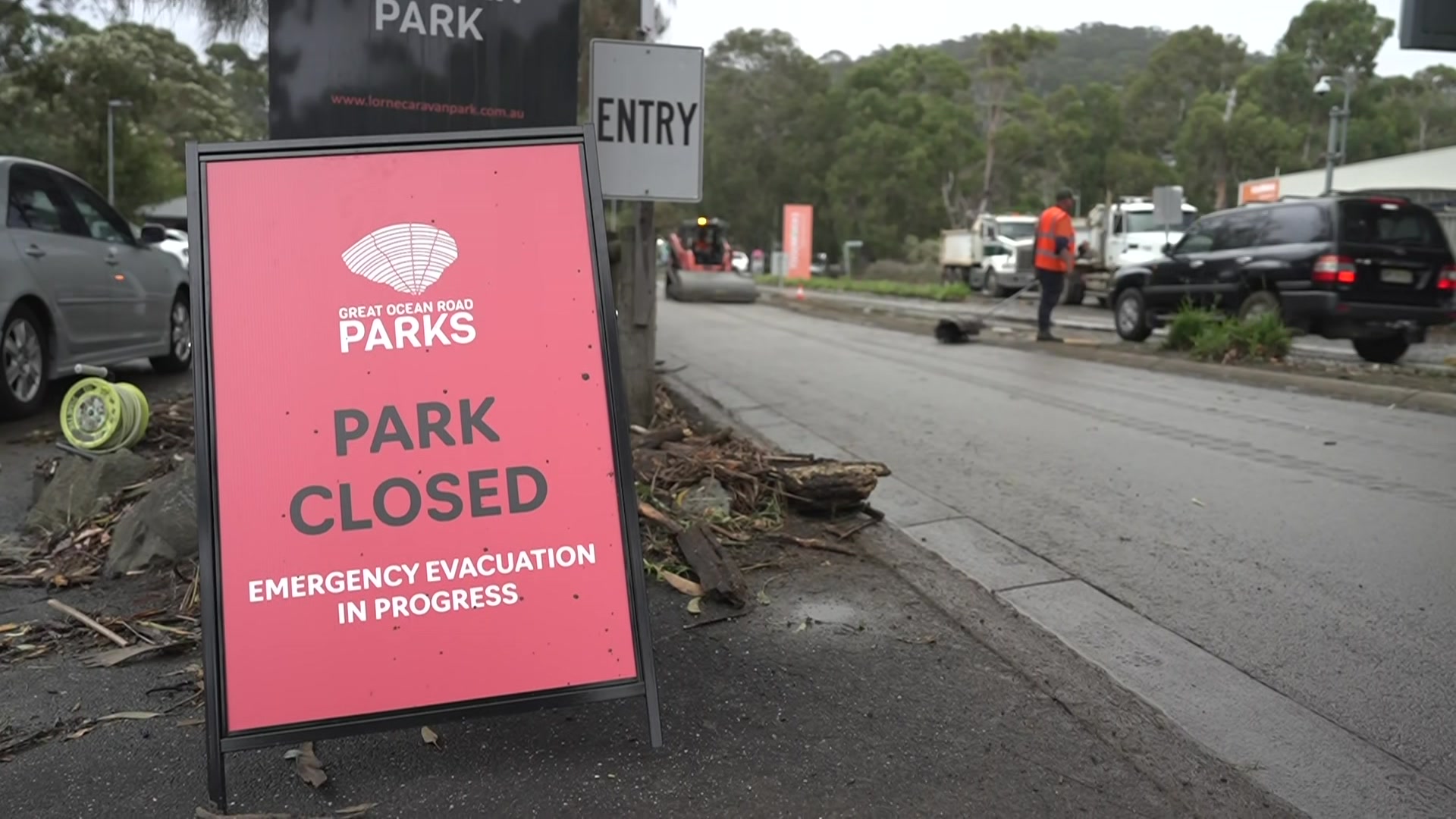 Uma placa vermelha em uma rodovia diz "parque fechado" e "evacuação de emergência em andamento" enquanto um homem de camisa laranja está perto de um carro.