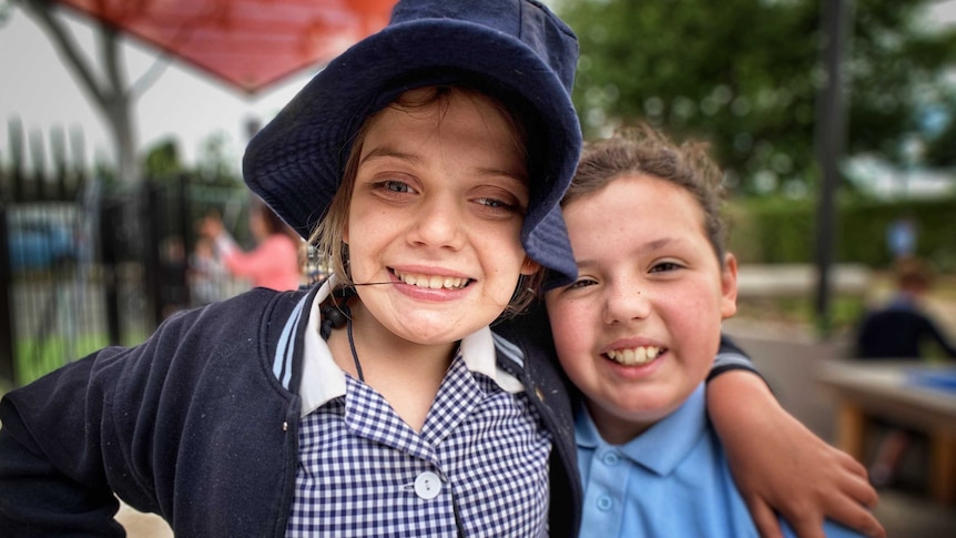 A picture of a girl in a school dress and hat with her arm around another girl in a school polo top.