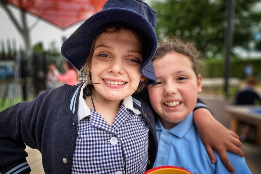 A picture of a girl in a school dress and hat with her arm around another girl in a school polo top.