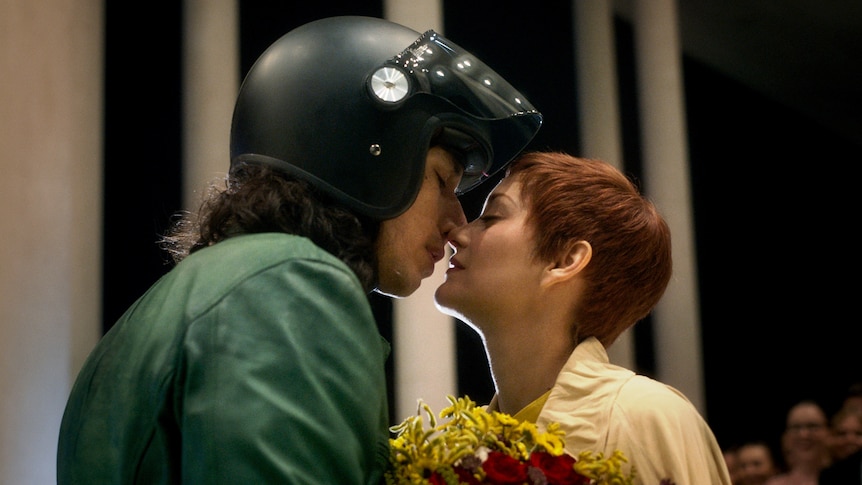 Adam Driver with lifted motorcycle helmet on the cusp of a tender kiss with Marion Cotillard who holds a bright floral bouquet.