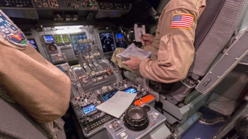 The central control panel of an 747, with pilots' arms bearing NASA insignia on their jumpsuits.