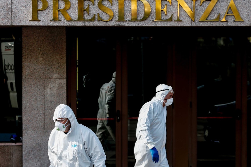 Two men in white overalls and face masks and blue gloves walk in front of a modern building.