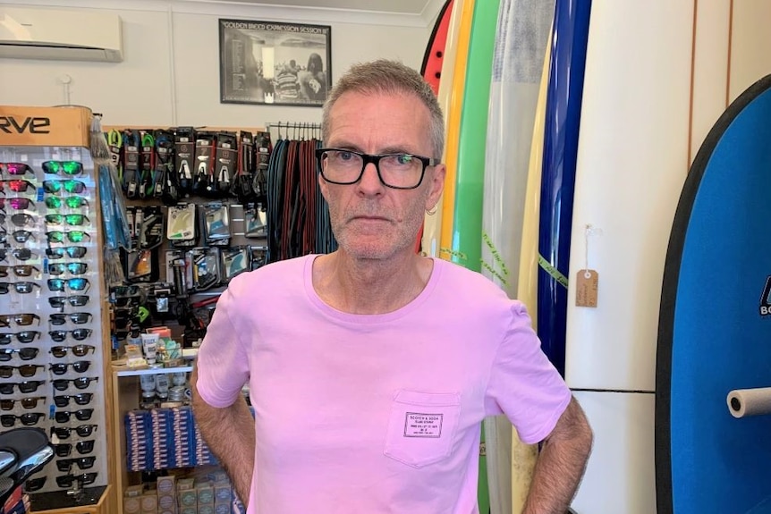 A man in black glasses and a purple t-shirt stands in a surf shop beside a row of surfboards.