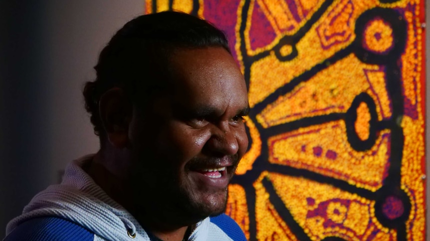 Tapaya Edwards smiles while sitting in front of an artwork from the Songlines exhibition.