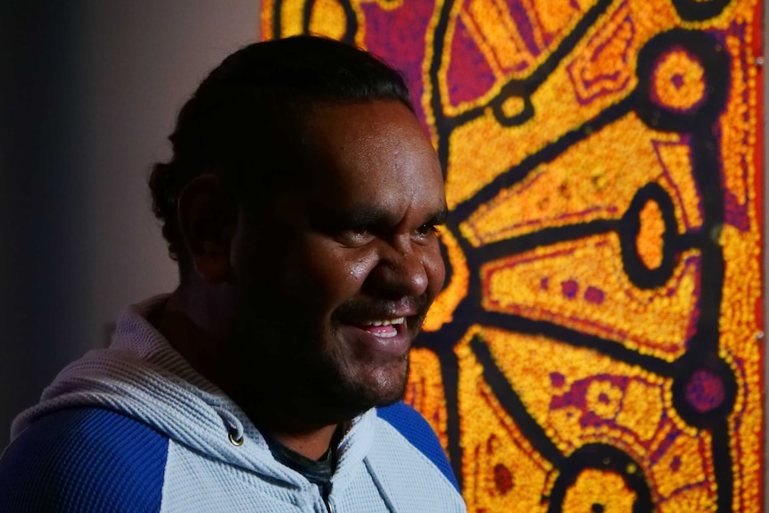 Tapaya Edwards smiles while sitting in front of an artwork from the Songlines exhibition.
