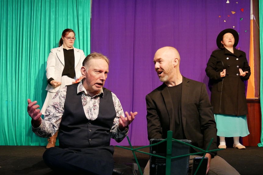 Two men sing together while sitting on a stage, two women dance in the background 