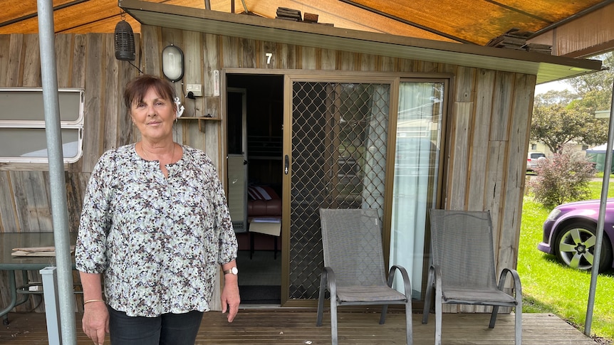 Angela wears a floral top and stands in front of her site which has wood paneling across the cabin and caravan 