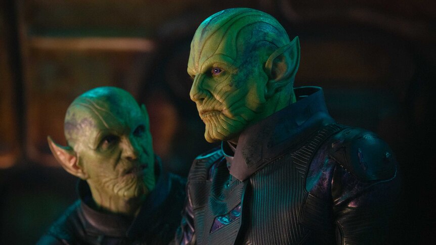 Actor Ben Mendelsohn stands in the foreground of the image in costume as a green, bald, alien with pointed ears and ridged skin.