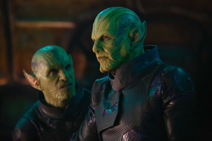 Actor Ben Mendelsohn stands in the foreground of the image in costume as a green, bald, alien with pointed ears and ridged skin.