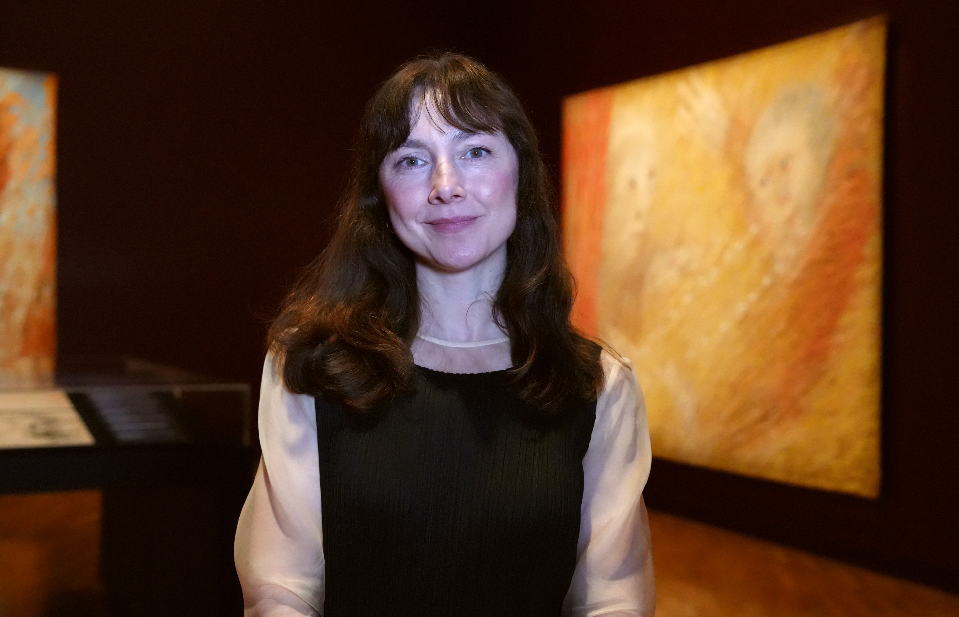 A woman with long drak hair stands in an art gallery smiling lightly.
