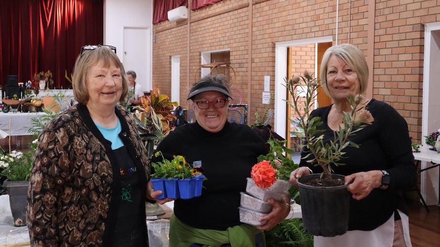 three women standing in a hall, holding flowers, and standing in front of a table of flowers.