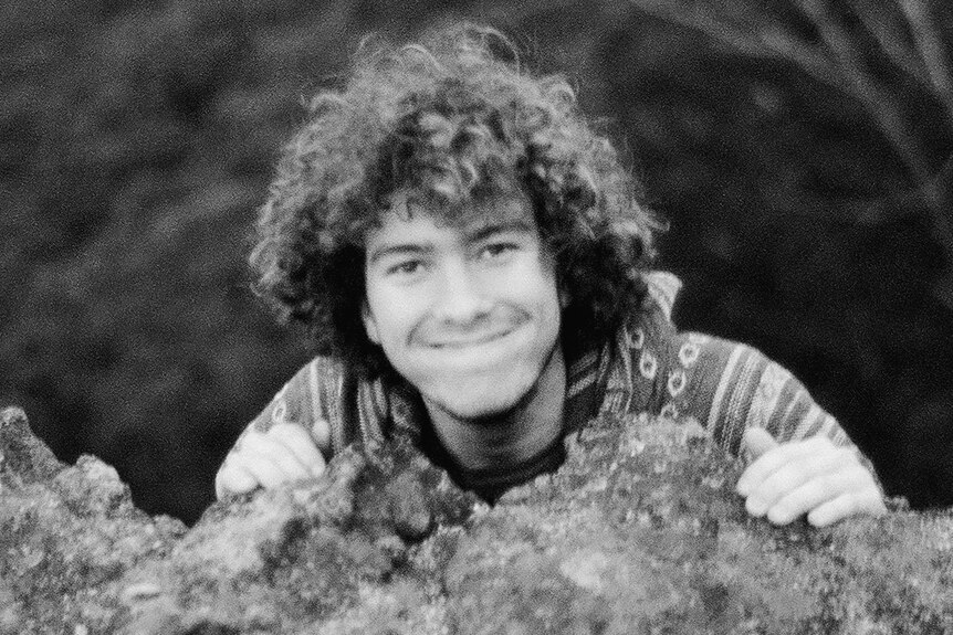 A young man smiling as he clings to the top of a rock