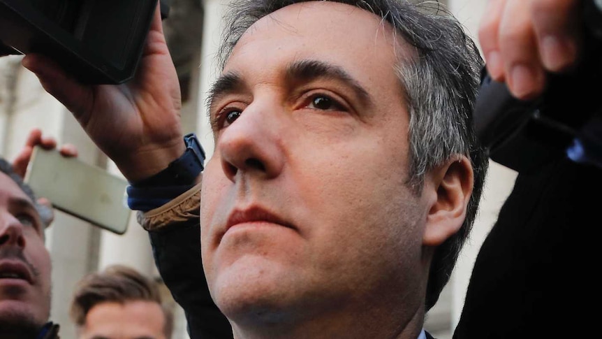 Michael Cohen, wearing a pale blue tie with his suit, is surrounded by journalists as he leaves court.