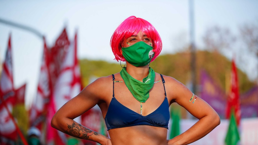 A woman wearing a pink wig and a green mask stand with her arms on her hips as people protest behind her