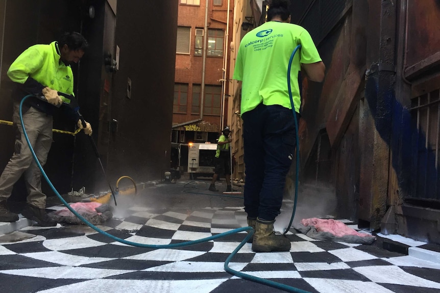 Two men wearing work boots and high visibility shirts use a pressure hose to remove painting from the road.