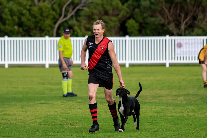 A man in football uniform smiles running a black dog from a football field 
