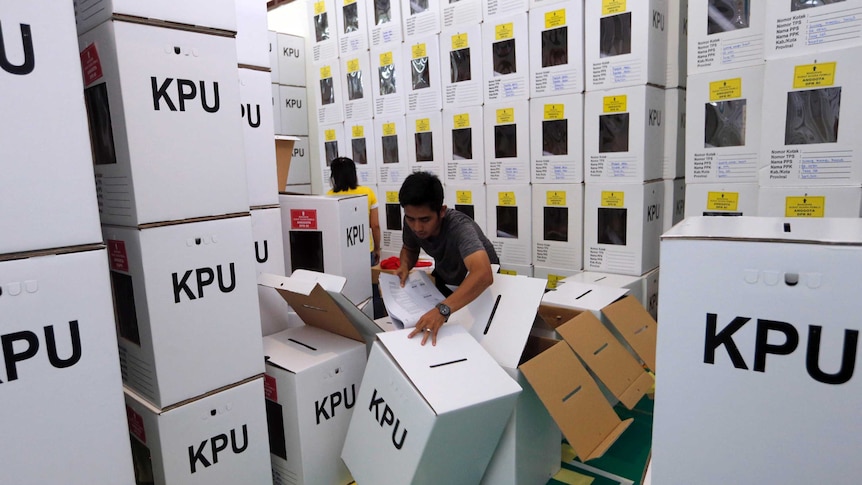 Stacks of Indonesian ballot boxes tower over a man who is struggling to grip one as it falls to the floor.