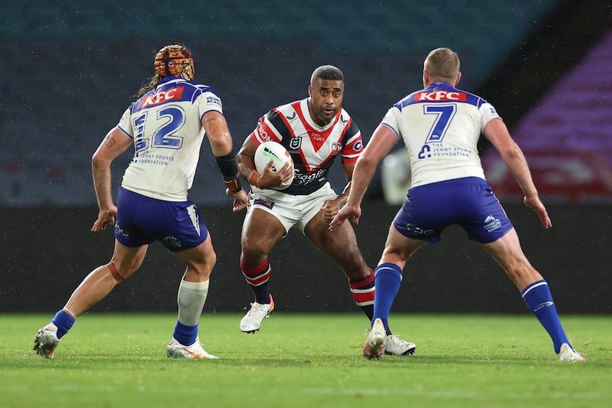 A rugby league player with ball in hand, running at a pair of defenders who brace for the tackle