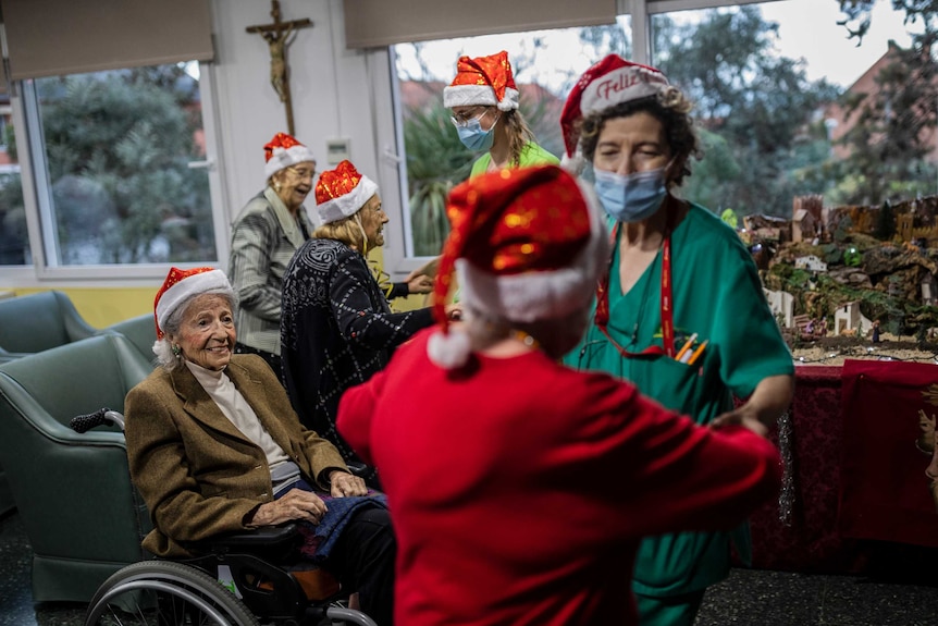 Elderly women wear Christmas hats and face masks while holding each other's hands.