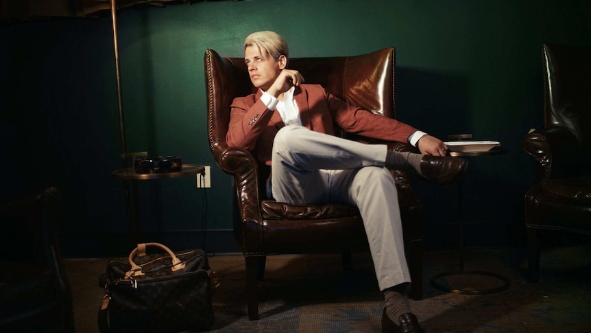 Milo Yiannopoulos sits in a brown leather chair, next to a large lamp.