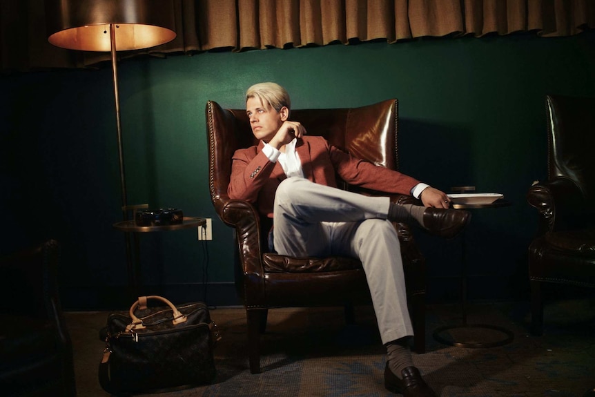 Milo Yiannopoulos sits in a brown leather chair, next to a large lamp.