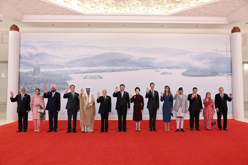 Ten men stand with four women all in a red carpet, waving.