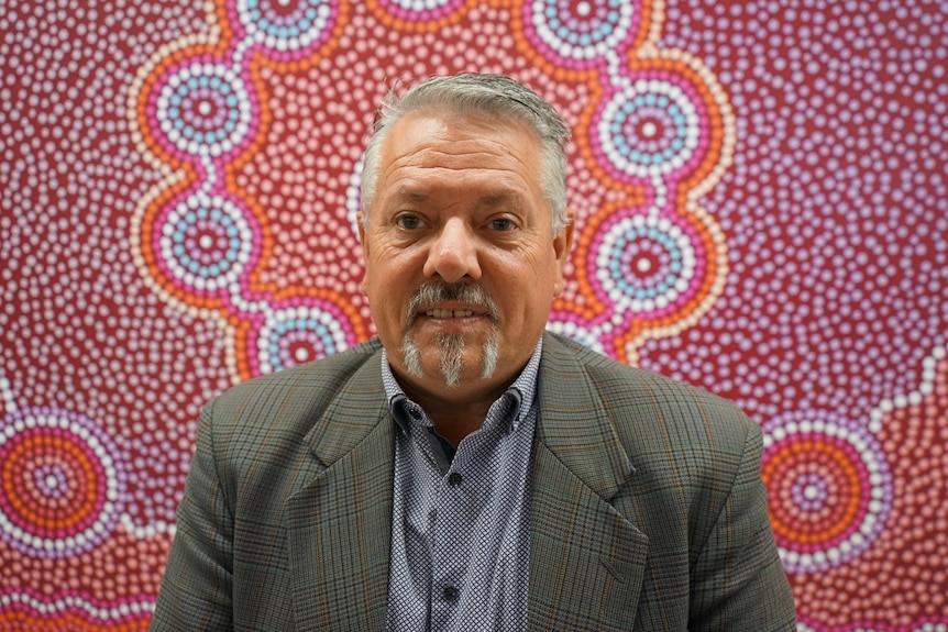 A grey-haired man stands in front an aboriginal painting