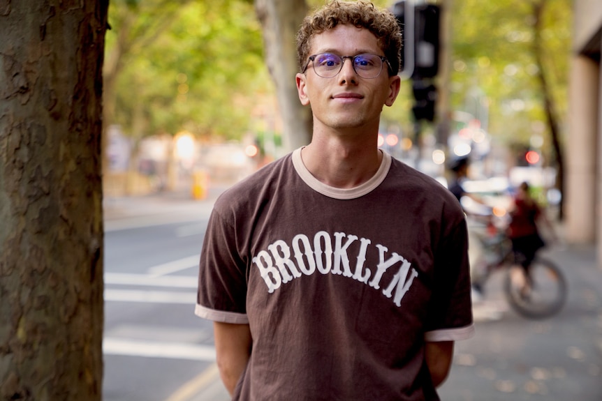 Young man with curly hair and glasses wearing brown t-shirt that reads 'Brooklyn'.