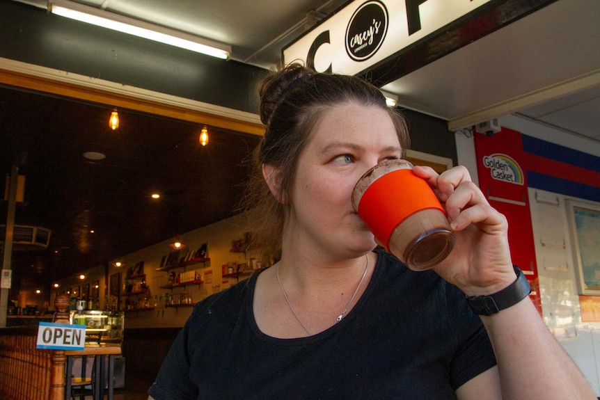 A woman in a black t-shirt sips a coffee from a reusable cup at an outside table outside a coffee shop on a town's main street.
