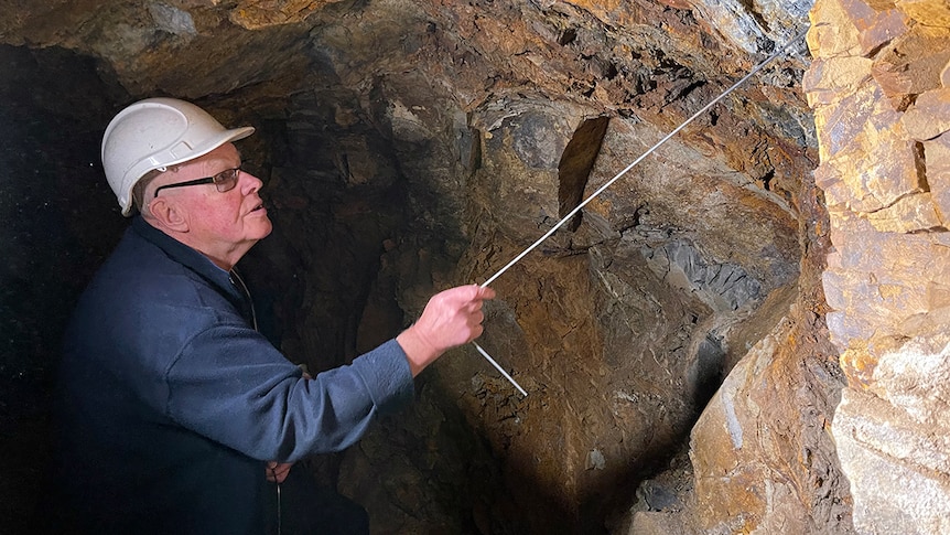 A man wearing a blue jacket and a white hard hat uses a metal rod to point at the rock wall of an underground mine shaft.
