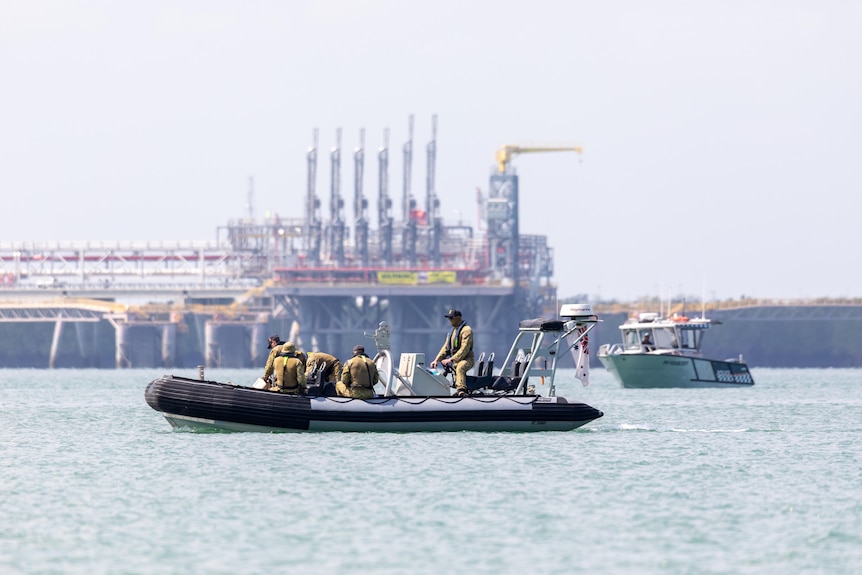A navy boat with four navy seamen on board crossing a harbour with a large industrial facility in the background.