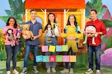 Five Play School presenters standing in a row, holding toys and smiling ion the brightly coloured Play School set.