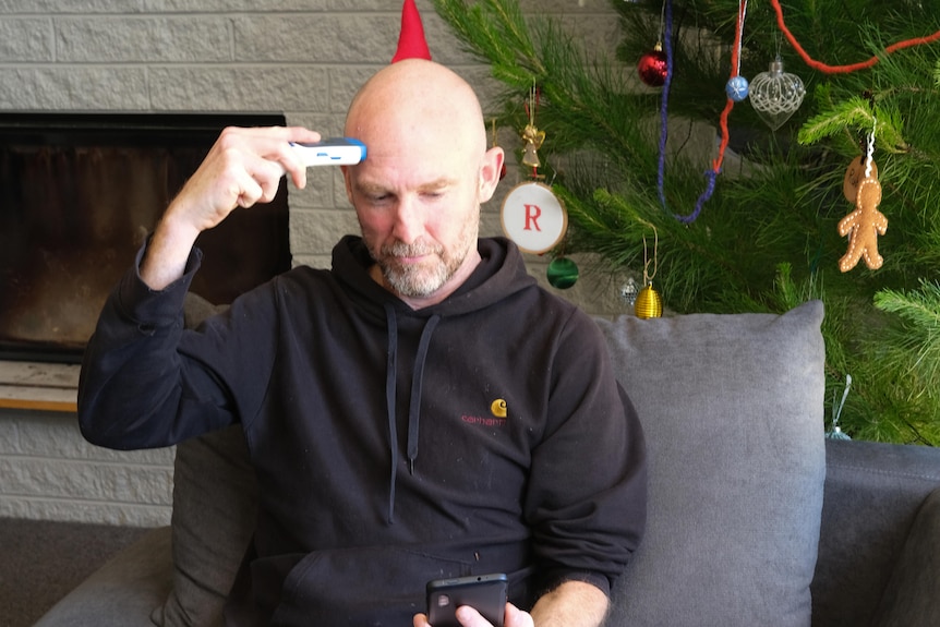 A man holds a thermometer to his forehead while looking down at his phone for results.