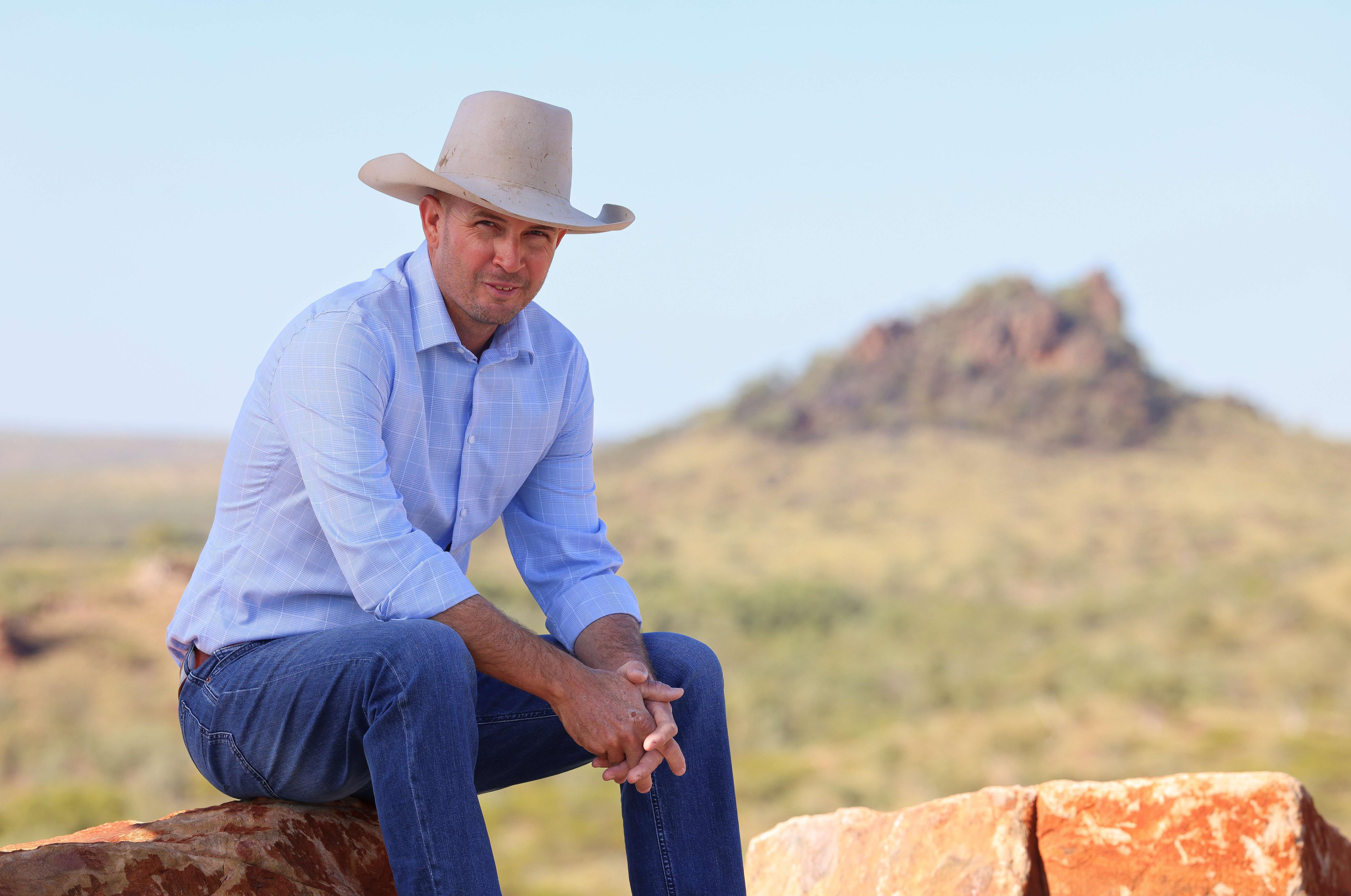 Um homem vestido com um terno do outback está sentado em uma rocha com uma paisagem rochosa atrás dele.