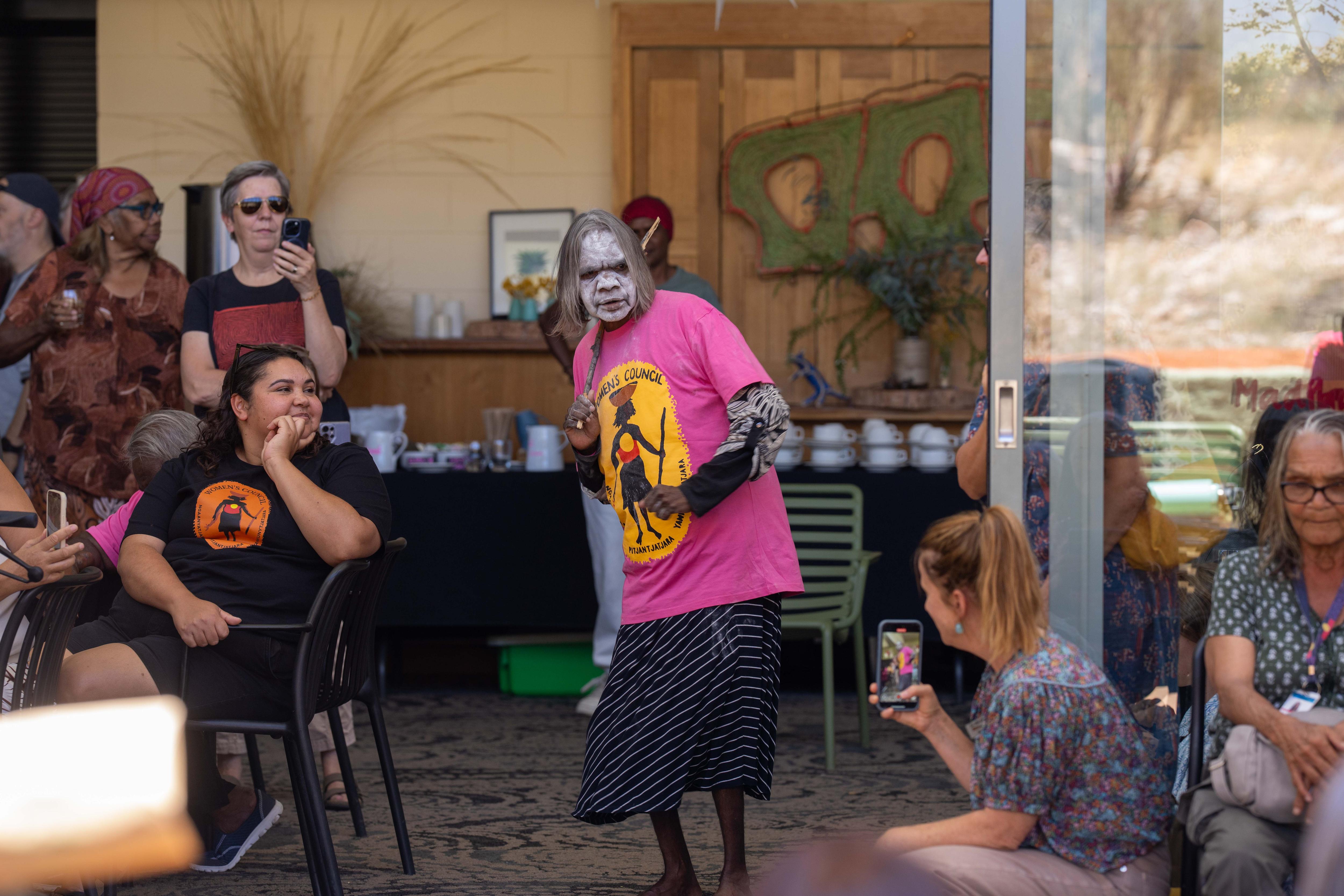 Una mujer indígena con el rostro pintado de blanco sostiene un palo y actúa frente a una multitud de espectadores.