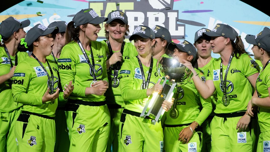 Sydney Thunder players smiles and stand around a large silver trophy in front of a colourful backdrop